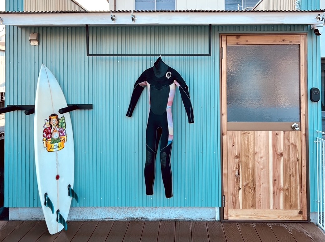 PRIVATE STAY HANARE — 徳島県海陽町宍喰浦松原 · ターコイズの外壁にサーフボードとウェットスーツを掛けた一棟貸しコテージの外観
