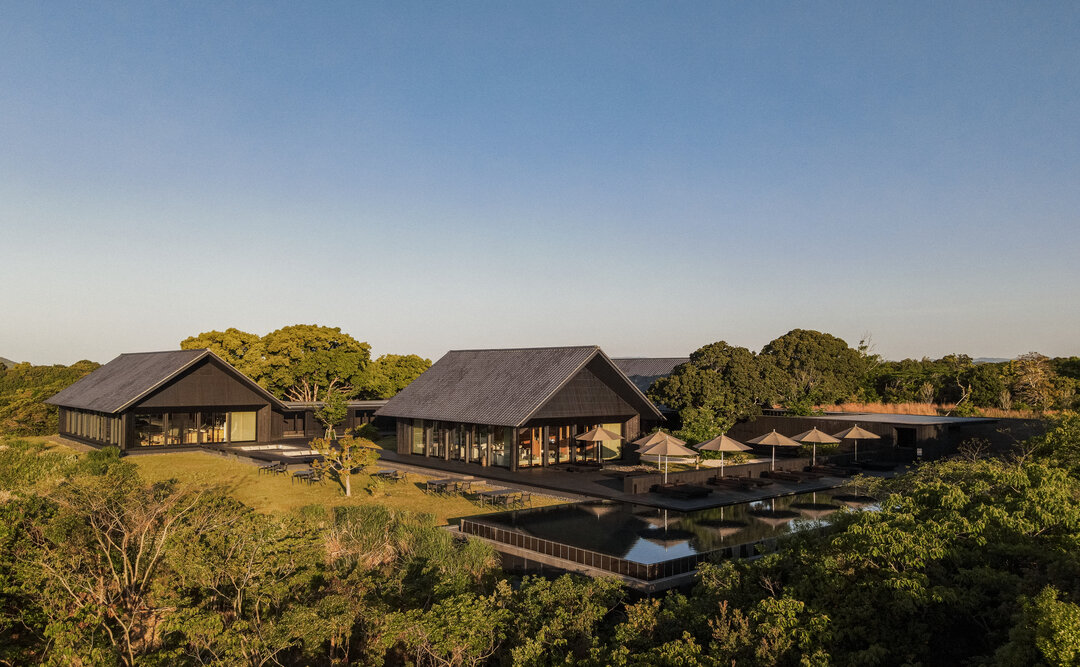 アマネム — 三重県志摩市浜島町 · 夕刻の光を受ける黒い切妻屋根の棟と、水面に棟を映す屋外プール