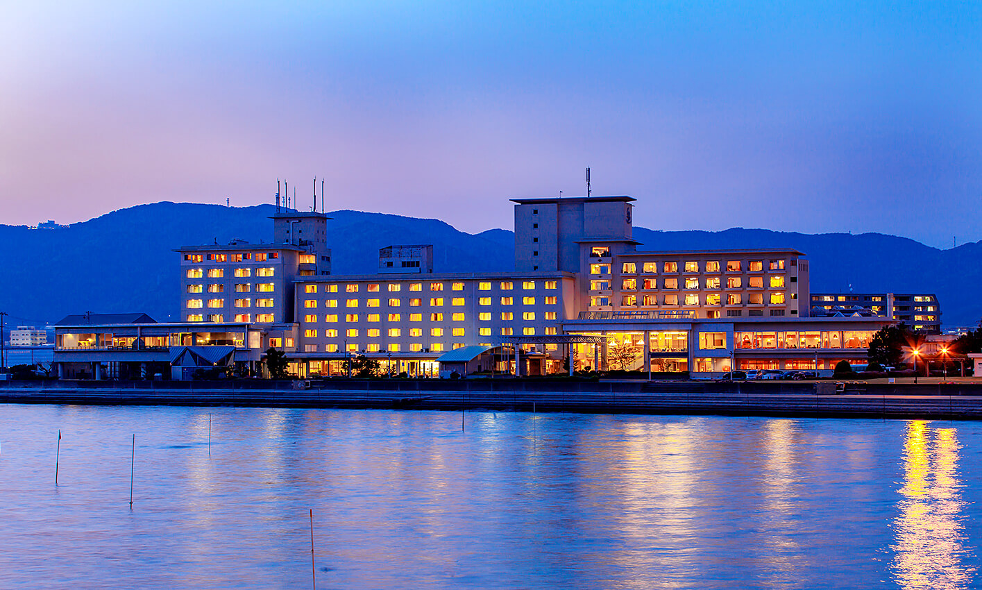 ホテル竹島 — 愛知・蒲郡竹島町 · 夕暮れの三河湾越しに灯りをともす海辺の外観