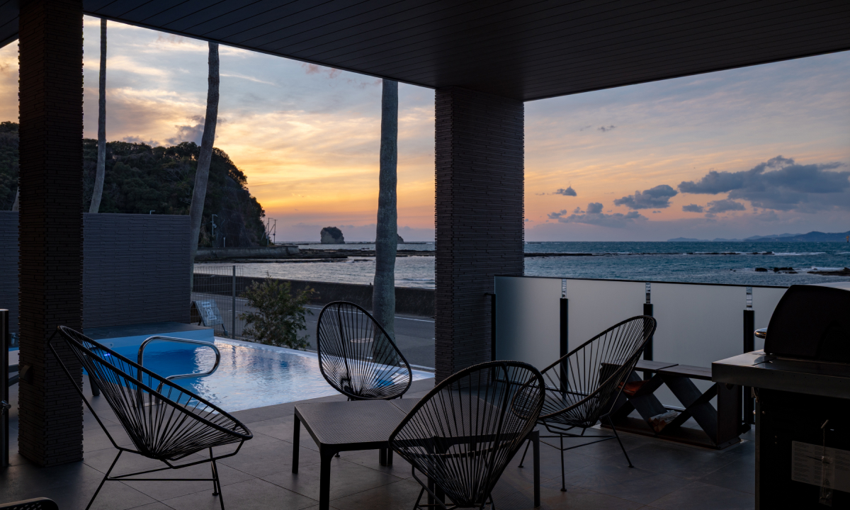 EZRA VILLA ~SHIRAHAMA~ — 和歌山県白浜町江津良 · 夕景の海に面した屋根付きテラスとインフィニティプール、薪を収めたラック